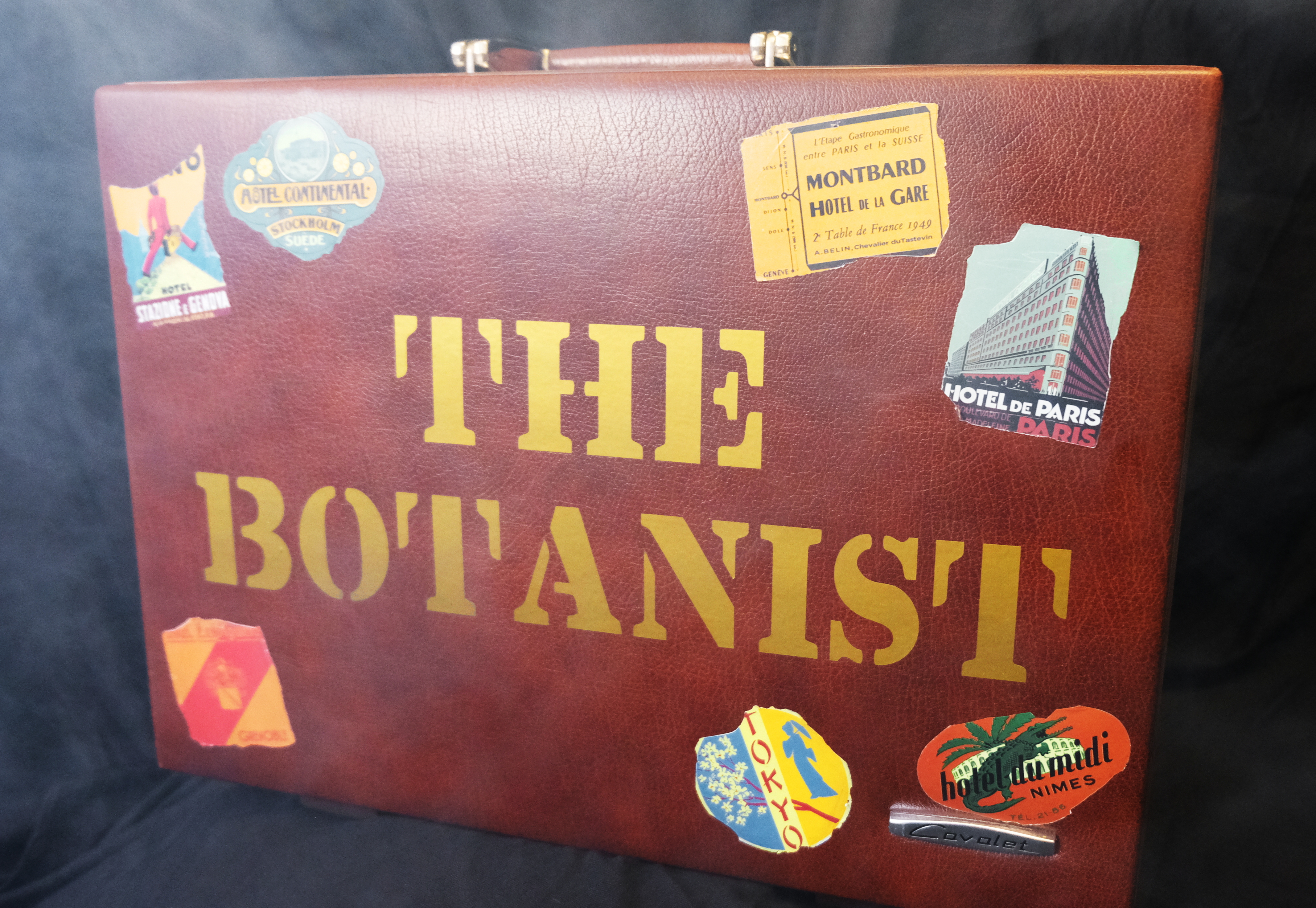 An old briefcase covered in tatty stickers from various cities across the world Large gold letters on the front read The Botanist